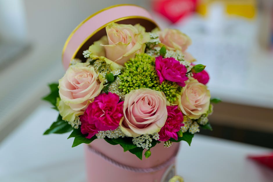 A round, pink floral gift box containing a fresh bouquet of pastel pink roses, bright pink carnations, and small green hypericum berries, accented with white baby's breath and lush green foliage, arra