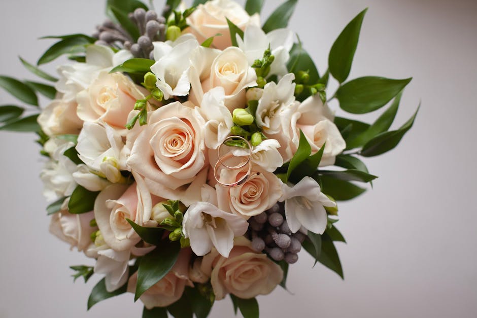 A close-up view of a floral bouquet arranged with pale peach and white roses, complemented by white freesias and clusters of small, round, lilac-colored berries. The arrangement is lush with vibrant g