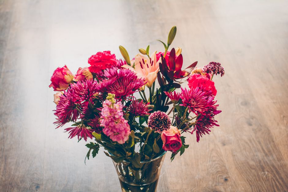 A vibrant bouquet arrangement of fresh flowers, including pink and red roses, purple chrysanthemums, peach lilies, and small pink statice, displayed in a clear glass vase on a wooden surface. The flow