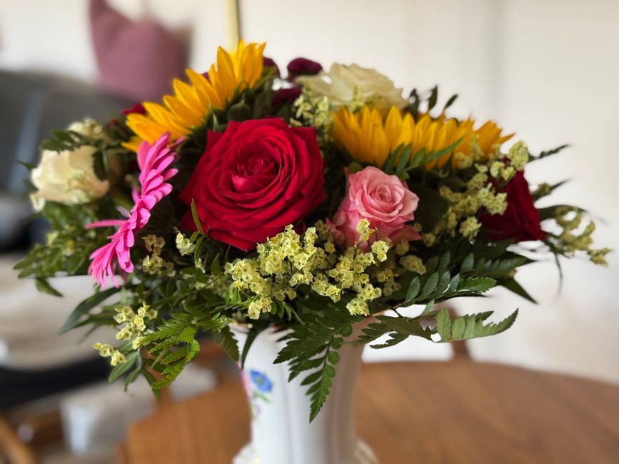 A vibrant floral arrangement in a white ceramic vase featuring a mix of fresh flowers including a large red rose, a soft pink rose, bright yellow sunflower-like blooms, and smaller white and pink blos