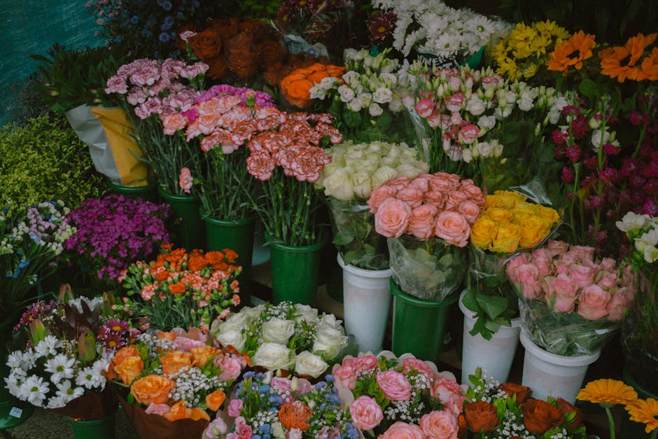 A vibrant display of fresh flowers arranged in various bouquets and individual stems, featuring shades of pink, white, yellow, orange, purple, and red. The flowers include roses, carnations, daisies, 