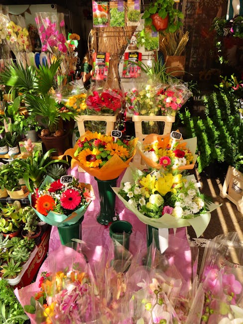 A display of fresh floral bouquets arranged on a pink cloth-covered surface inside a flower shop, with vibrant flowers including red, yellow, pink, and white blooms. The bouquets are wrapped in colorf
